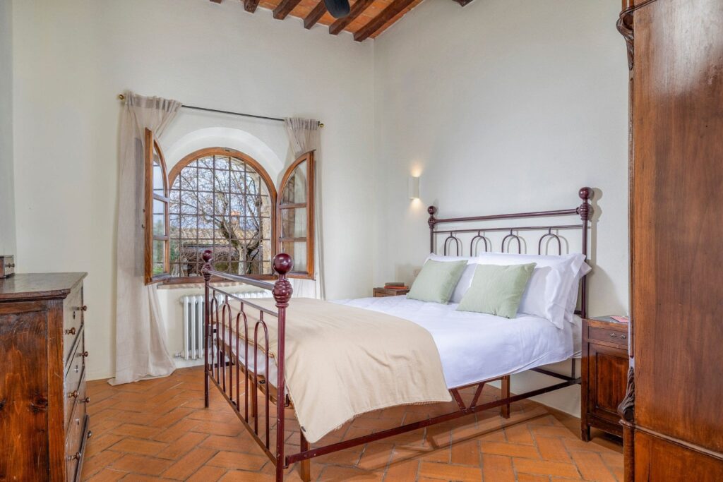 Camera matrimoniale dell’appartamento Orcia con letto in ferro battuto, travi in legno e finestra ad arco affacciata sulla campagna toscana